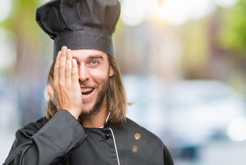 Young handsome cook man with long hair over isolated background covering one eye with hand with confident smile on face and surprise emotion.