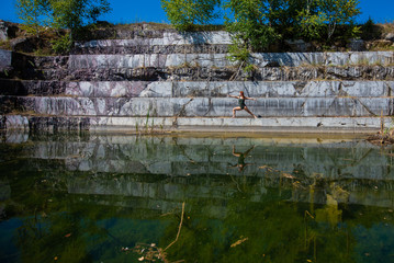 Fototapeta na wymiar A red-haired young woman in green overalls is engaged in yoga at the degree of an abandoned marble quarry. In the water of a flooded marble quarry the reflection of a girl engaged in pilates