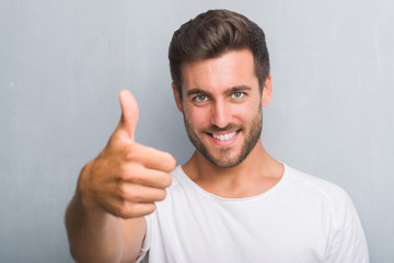 Handsome young man over grey grunge wall happy with big smile doing ok sign, thumb up with fingers, excellent sign