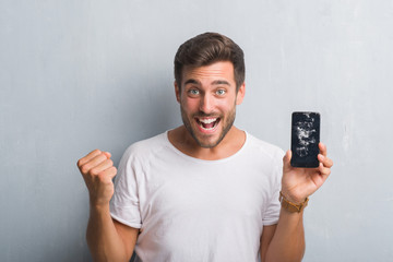 Handsome young man over grey grunge wall showing broken smartphone screen screaming proud and celebrating victory and success very excited, cheering emotion