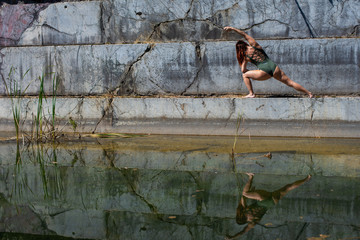 A red-haired young woman in green overalls is engaged in yoga at the degree of an abandoned marble quarry. In the water of a flooded marble quarry the reflection of a girl engaged in pilates