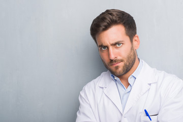 Handsome young professional man over grey grunge wall wearing white coat skeptic and nervous, disapproving expression on face with crossed arms. Negative person.