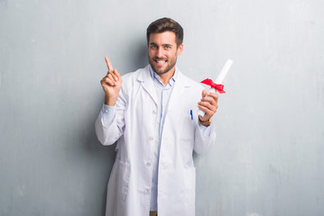 Handsome young doctor man over grey grunge wall holding diploma very happy pointing with hand and...