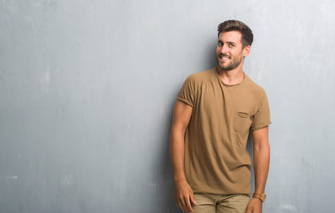Handsome young man over grey grunge wall looking away to side with smile on face, natural expression. Laughing confident.