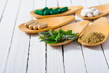 herbal leaves, ground herb powder and medicament pills on bamboo spoons, white wooden table