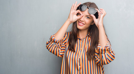 Young brunette woman over grunge grey wall wearing big sunglasses with happy face smiling doing ok sign with hand on eye looking through fingers