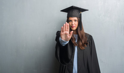 Young brunette woman over grunge grey wall wearing graduate uniform with open hand doing stop sign with serious and confident expression, defense gesture