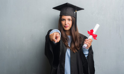 Young brunette woman over grunge grey wall wearing graduate uniform holding degree pointing with finger to the camera and to you, hand sign, positive and confident gesture from the front