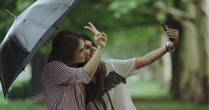 Closeup in the middle of the parc two ladies taking selfies under a black umbrella.