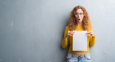 Young redhead woman over grey grunge wall holding blank paper sheet scared in shock with a surprise face, afraid and excited with fear expression