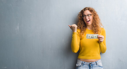 Young redhead woman over grey grunge wall holding a dollar pointing and showing with thumb up to the side with happy face smiling