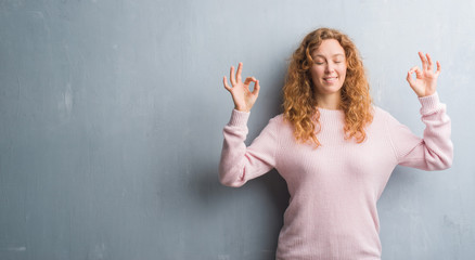 Young redhead woman over grey grunge wall wearing pink sweater relax and smiling with eyes closed doing meditation gesture with fingers. Yoga concept.