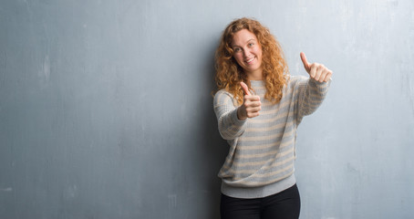 Young redhead woman over grey grunge wall approving doing positive gesture with hand, thumbs up smiling and happy for success. Looking at the camera, winner gesture.