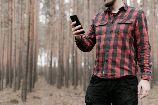 Man in red plaid shirt and black military pants holds a smartphone in his hand. Lost in the pine forest. Copy space. Toned