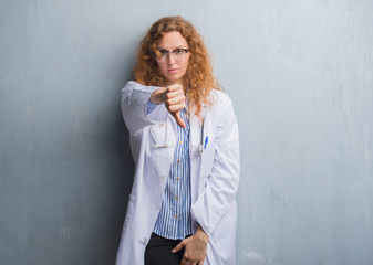 Young redhead doctor woman over grey grunge wall wearing a coat looking unhappy and angry showing rejection and negative with thumbs down gesture. Bad expression.