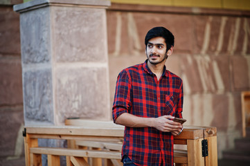 Young indian student man at red checkered shirt and jeans posed at city with mobile phone at hands.