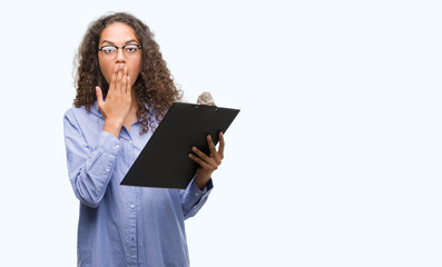 Young hispanic business woman holding clipboard cover mouth with hand shocked with shame for mistake, expression of fear, scared in silence, secret concept