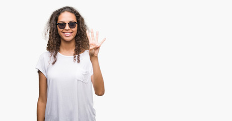 Young hispanic woman wearing sunglasses showing and pointing up with fingers number four while smiling confident and happy.