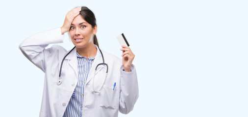 Young hispanic doctor woman holding credit card stressed with hand on head, shocked with shame and surprise face, angry and frustrated. Fear and upset for mistake.