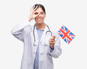 Young hispanic doctor woman holding flag of united kingdom with happy face smiling doing ok sign with hand on eye looking through fingers