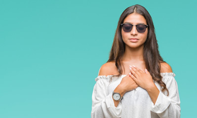 Young beautiful arab woman wearing sunglasses over isolated background smiling with hands on chest with closed eyes and grateful gesture on face. Health concept.
