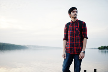 Young indian student man at checkered shirt and jeans with backpack posed on evening city against lake.