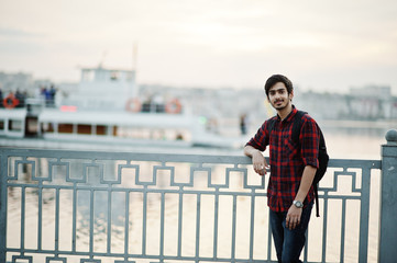 Young indian student man at checkered shirt and jeans with backpack posed on evening city against lake and boat.