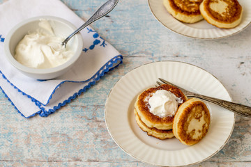 Delicious fried syrniki on a round plate, next to a sour cream cup on a cotton napkin on an old wooden table. 