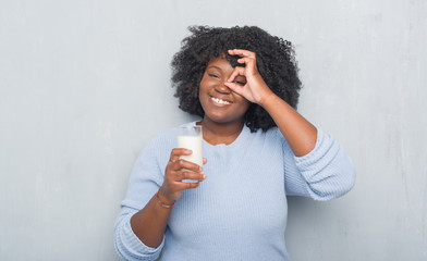 Young african american woman over grey grunge wall drinking a glass of milk with happy face smiling doing ok sign with hand on eye looking through fingers