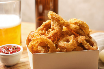 Cardboard box with crunchy fried onion rings and tomato sauce on wooden board, closeup