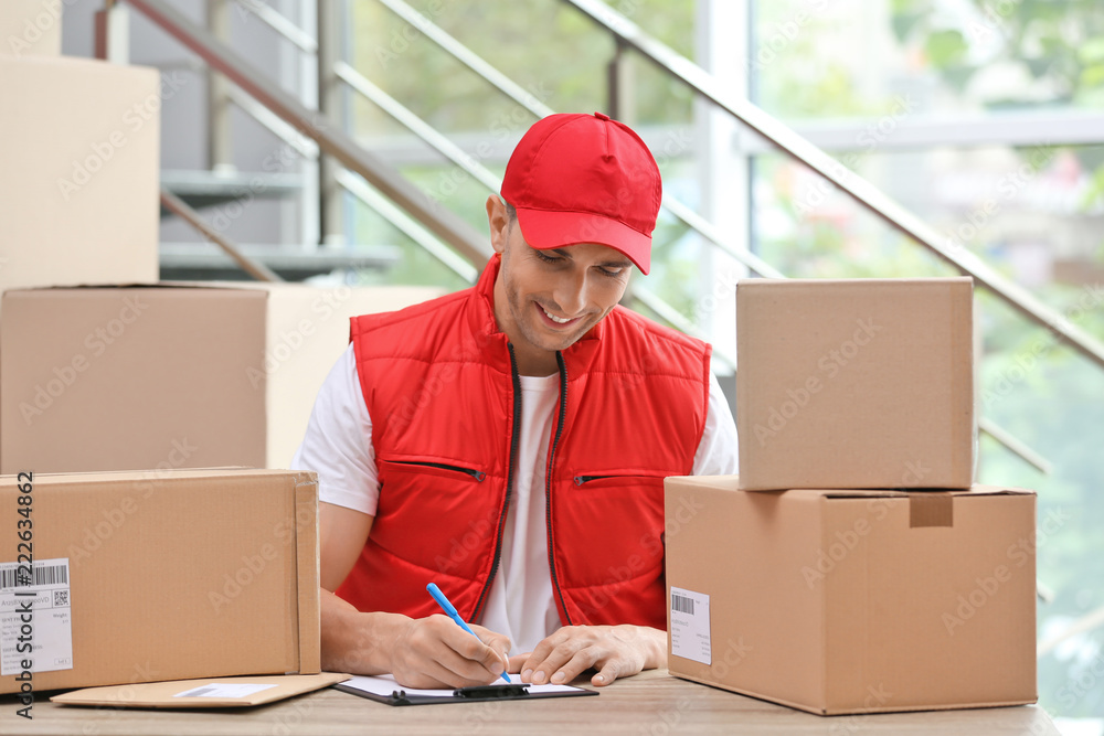 Sticker Young courier working with papers among parcels at table in delivery department