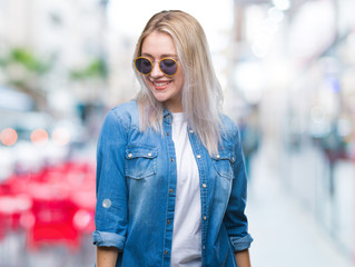 Young blonde woman wearing sunglasses over isolated background looking away to side with smile on face, natural expression. Laughing confident.