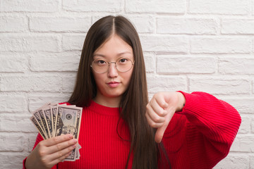 Young Chinese woman over brick wall holding dollars with angry face, negative sign showing dislike...