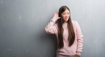 Young Chinese woman over grey wall smiling with hand over ear listening an hearing to rumor or gossip. Deafness concept.