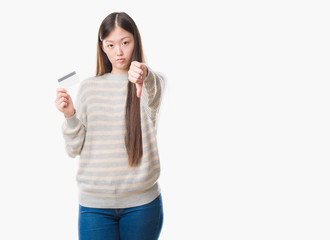 Young Chinese woman over isolated background holding credit card with angry face, negative sign showing dislike with thumbs down, rejection concept