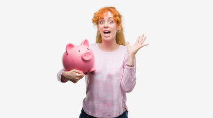 Young redhead woman holding piggy bank very happy and excited, winner expression celebrating victory screaming with big smile and raised hands