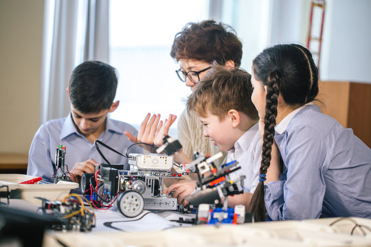 Never getting bored together. Adorable youngsters sitting at a table with their teacher and learning programming process of their new robot.