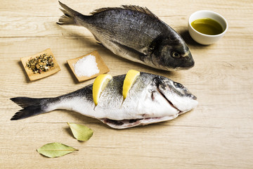 Two fresh fish Dorada with bay leaves, some pieces of lemon, a bowl of oil and some spices on a wooden table prepared for cooking
