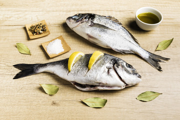 Two fresh fish Dorada with bay leaves, some pieces of lemon, a bowl of oil and some spices on a wooden table prepared for cooking
