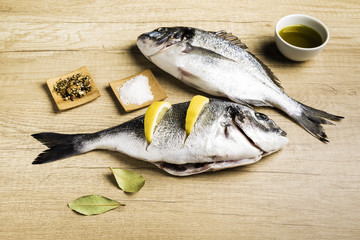 Two fresh fish Dorada with bay leaves, some pieces of lemon, a bowl of oil and some spices on a wooden table prepared for cooking
