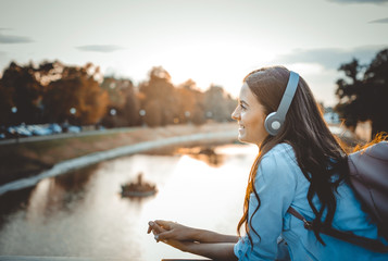 Happy young girl student listening to music while standing on a bridge at sunset. Smiling and looking away. - Powered by Adobe