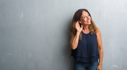 Middle age hispanic woman standing over grey grunge wall smiling with hand over ear listening an...