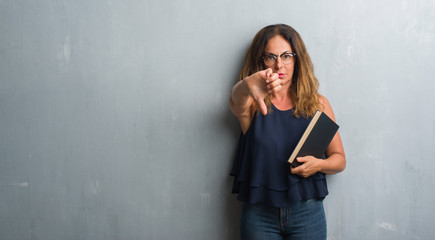 Middle age hispanic woman standing over grey grunge wall holding a book with angry face, negative...