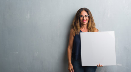 Middle age hispanic woman standing over grey grunge wall holding blank banner with a happy face standing and smiling with a confident smile showing teeth