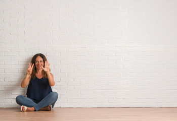 Middle age hispanic woman sitting on the floor over white brick wall showing and pointing up with fingers number eight while smiling confident and happy.