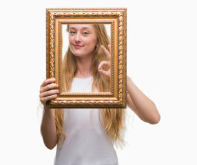 Blonde teenager woman holding vintage frame art doing ok sign with fingers, excellent symbol