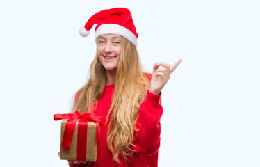 Blonde teenager woman wearing santa claus hat very happy pointing with hand and finger to the side