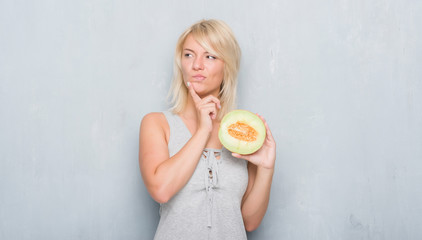 Adult caucasian woman over grunge grey wall eating cantaloupe melon serious face thinking about question, very confused idea