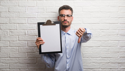 Young adult business man over brick wall holding clipboard with angry face, negative sign showing...