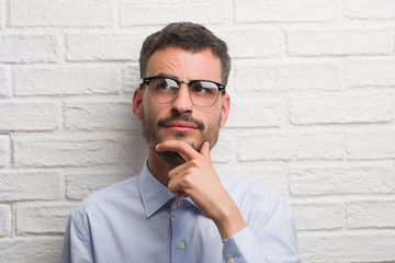 Young adult business man standing over white brick wall serious face thinking about question, very...